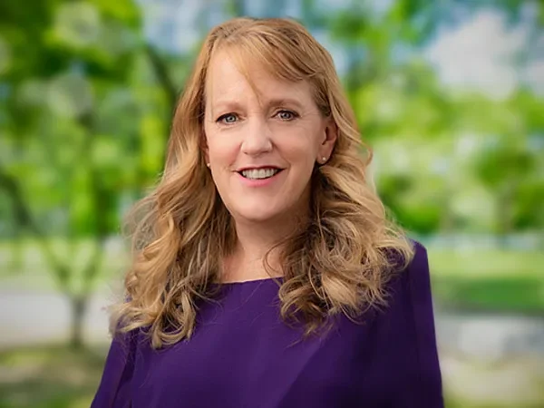 A woman with blonde curly hair smiling outdoors in a purple top, with a blurred green park background.