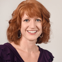 Portrait of a woman with red hair, wearing a purple top and earrings, smiling against a light background.