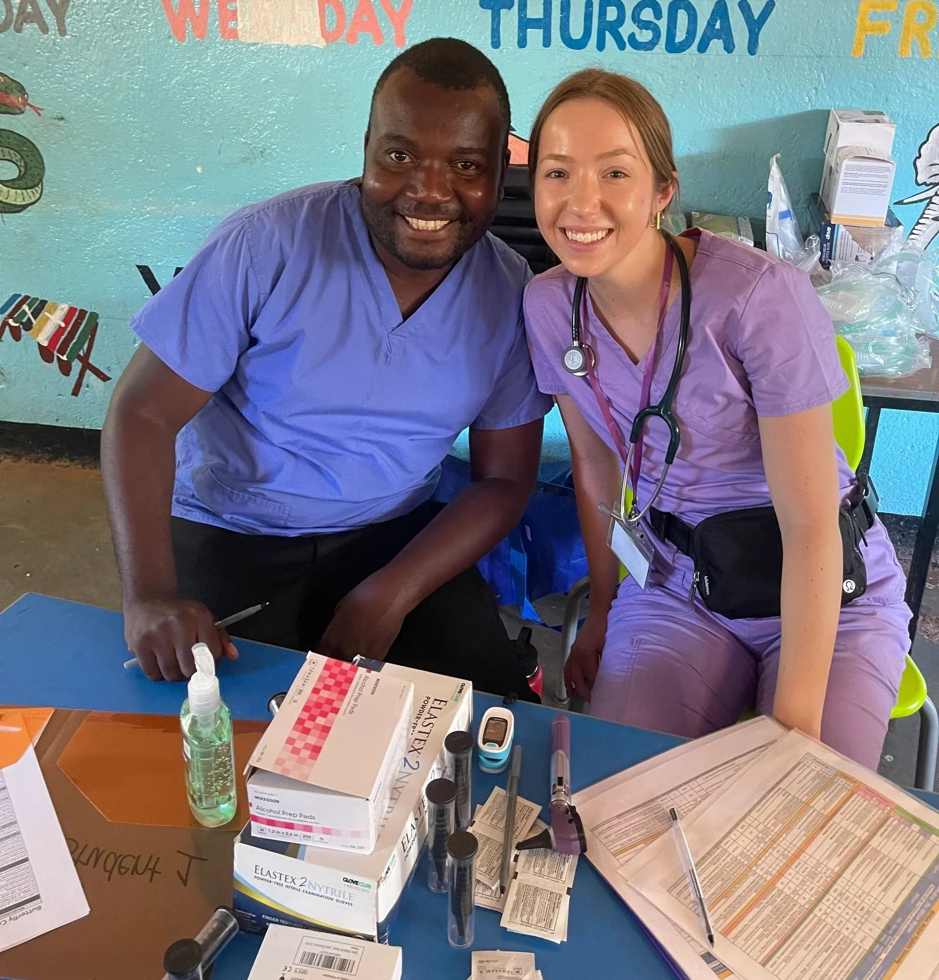 Two healthcare workers, a man and a woman, smiling at the camera in a clinical setting. The man is wearing blue scrubs and the woman is wearing purple scrubs with a stethoscope around her neck. They are seated at a table with medical supplies, includ