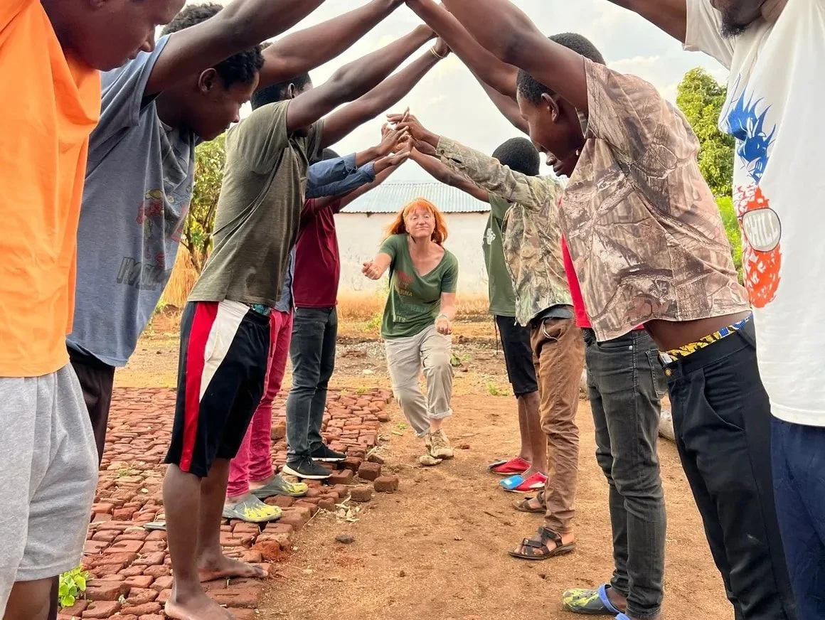 A woman with red hair walking through an arched tunnel of hands created by a group of children standing in two lines on either side of a dirt path, outdoors.