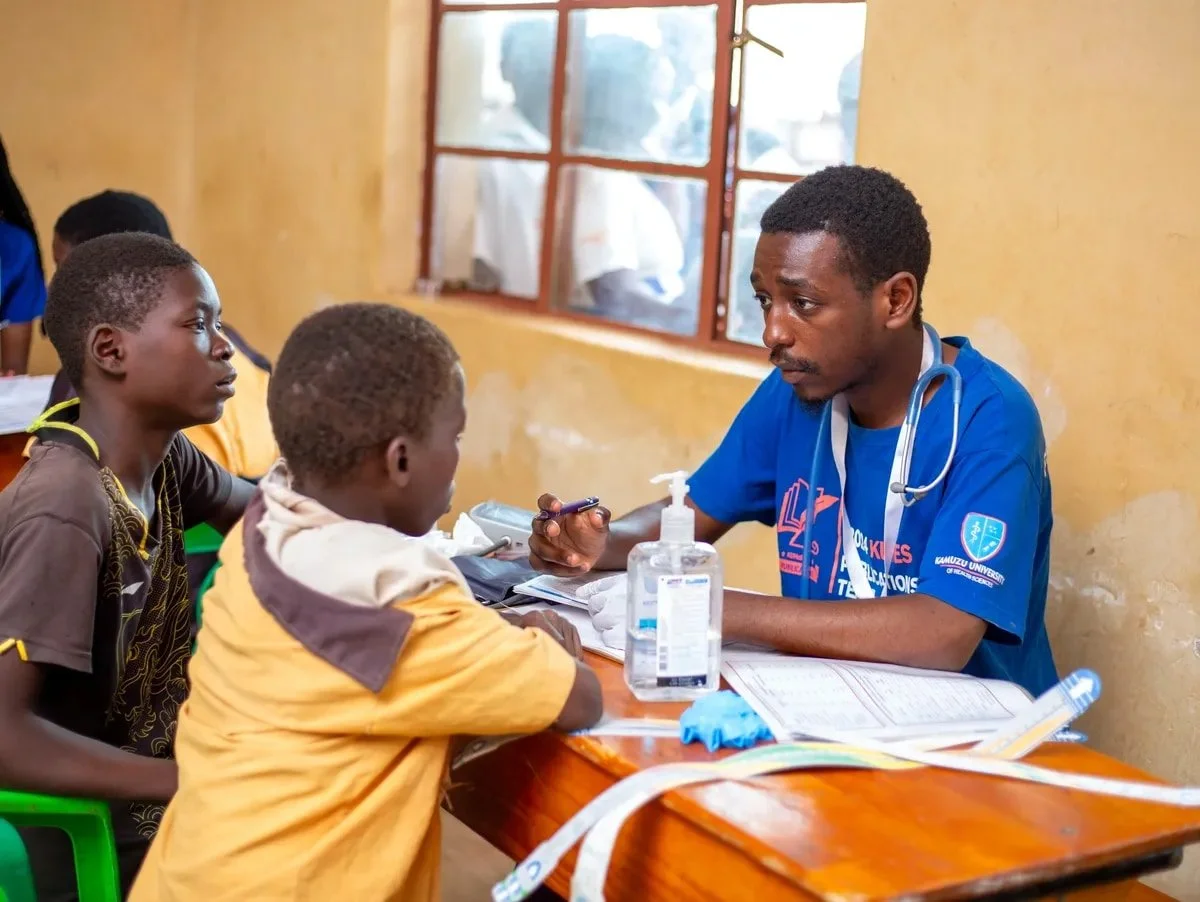 A healthcare professional consulting with two young children in a clinic, sitting at a wooden table with medical supplies, including hand sanitizer and papers.