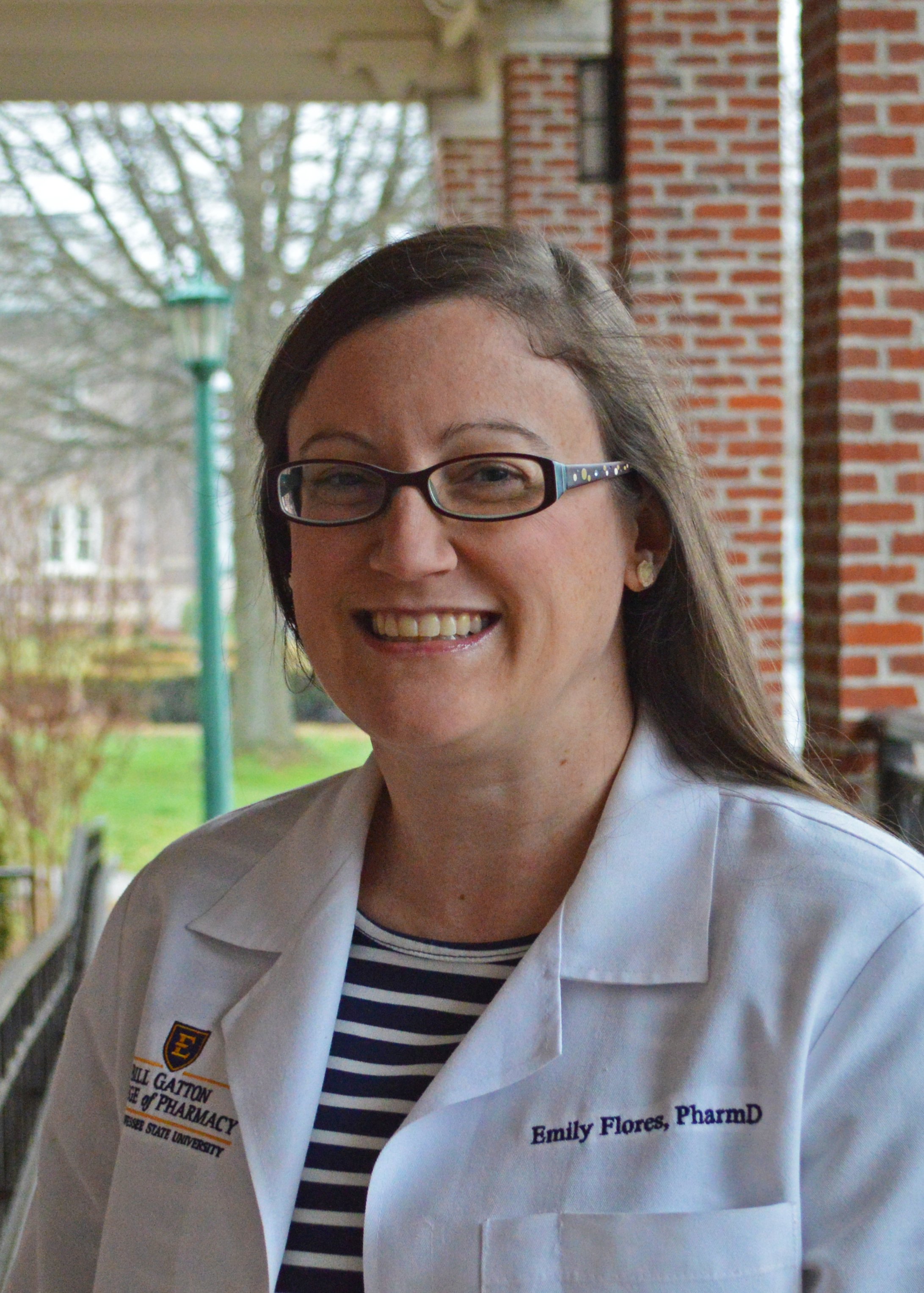 A woman smiling outdoors, wearing glasses, a striped navy and white shirt, and a white coat with embroidered text, near a brick building, trees, and a green lamppost.