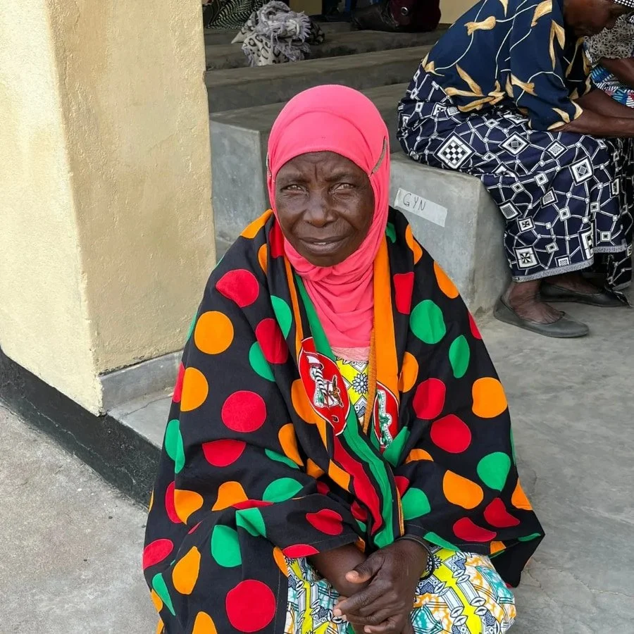 An elderly woman wearing a pink headscarf and a black shawl with large red, green, and orange polka dots, sitting on the ground outdoors, with another person sitting nearby.