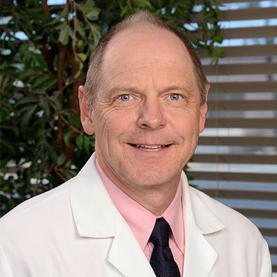 A middle-aged man with short, light hair, wearing a white medical coat over a pink shirt and navy tie, standing in front of a plant and window blinds.