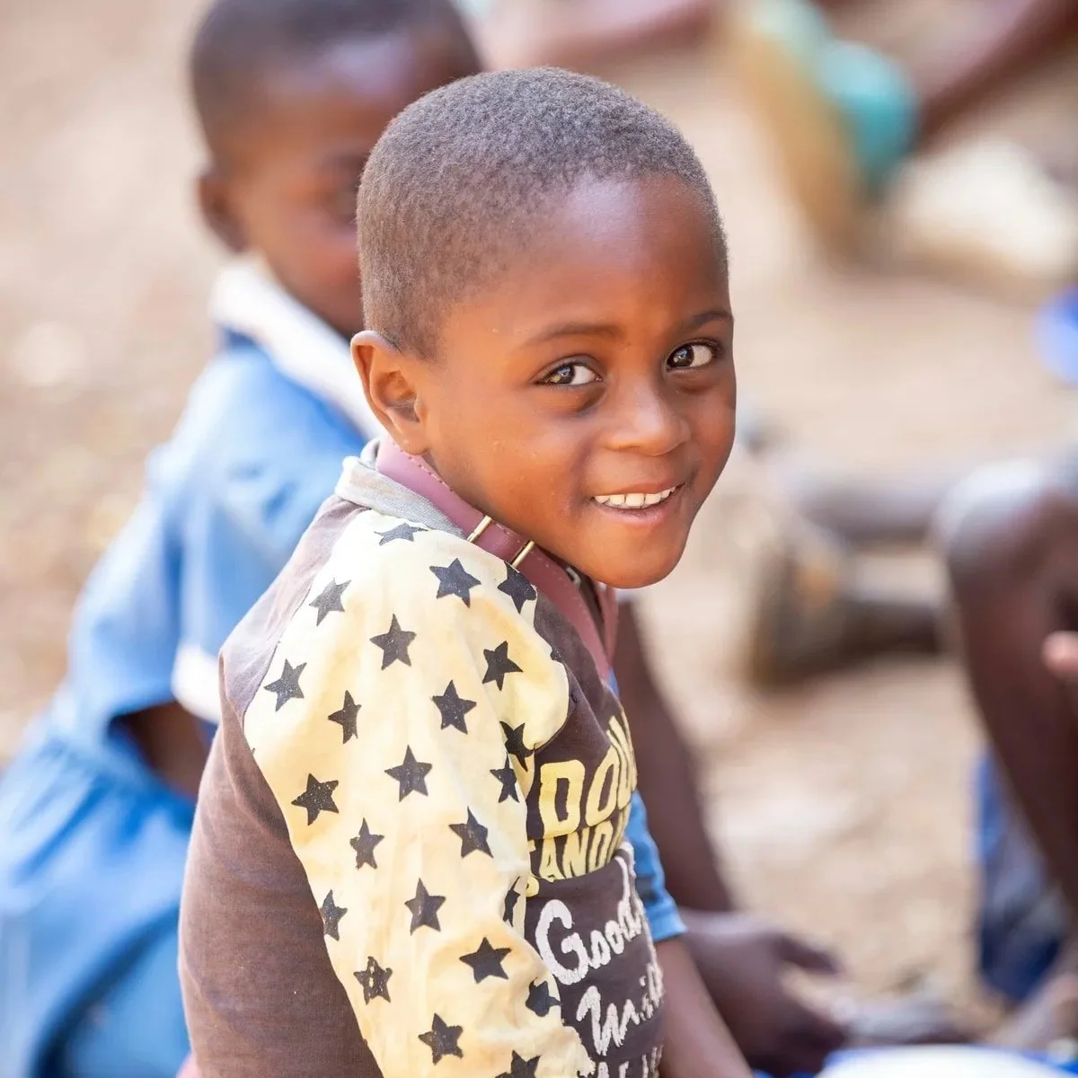 A young boy with a shaved head wearing a brown T-shirt with yellow sleeves and black star patterns, smiling at the camera, with other children blurred in the background.
