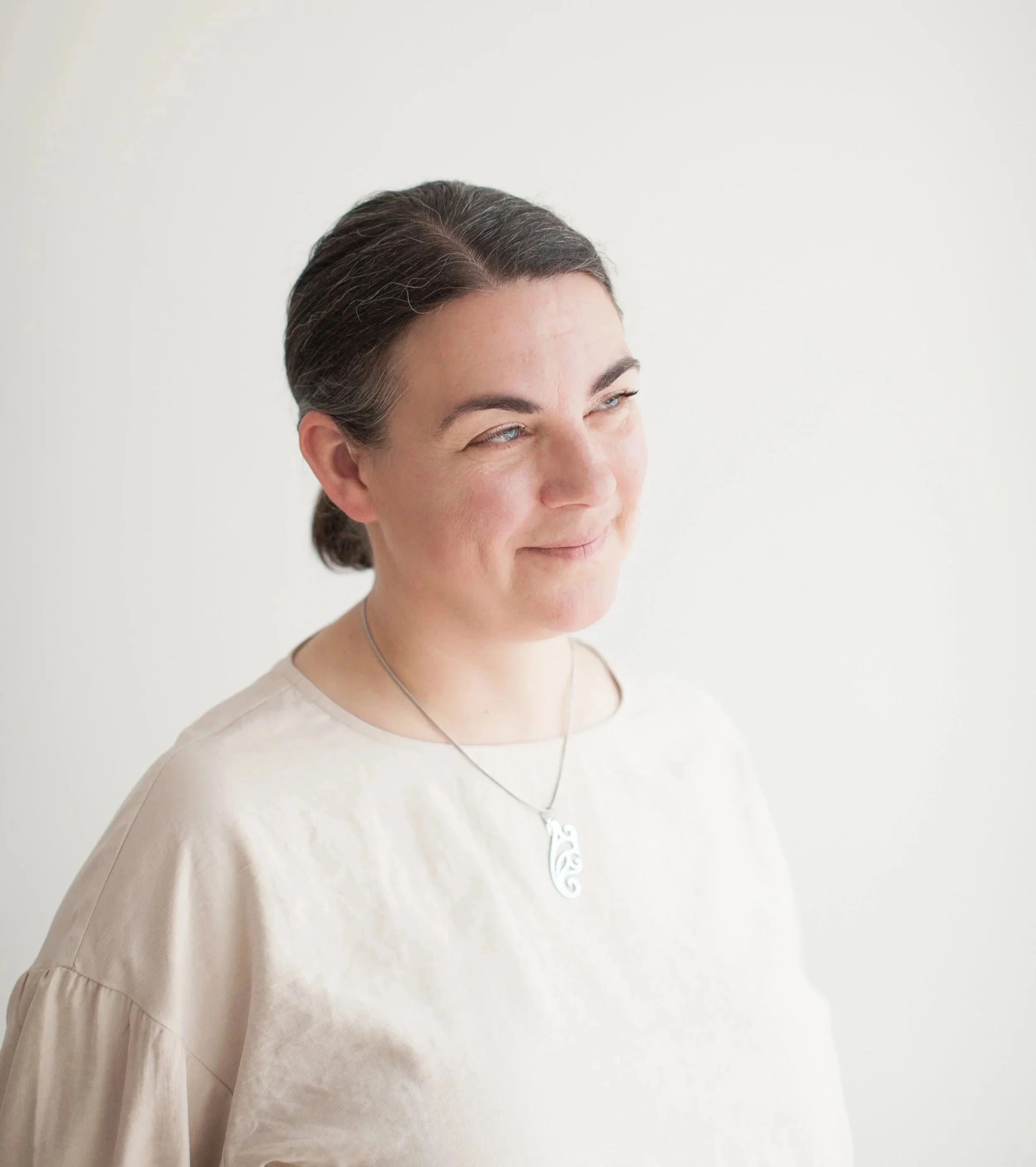 A woman with dark hair pulled back, wearing a cream-colored top with puffed sleeves, smiles slightly, wearing a silver necklace with a pendant, standing against a plain white background.