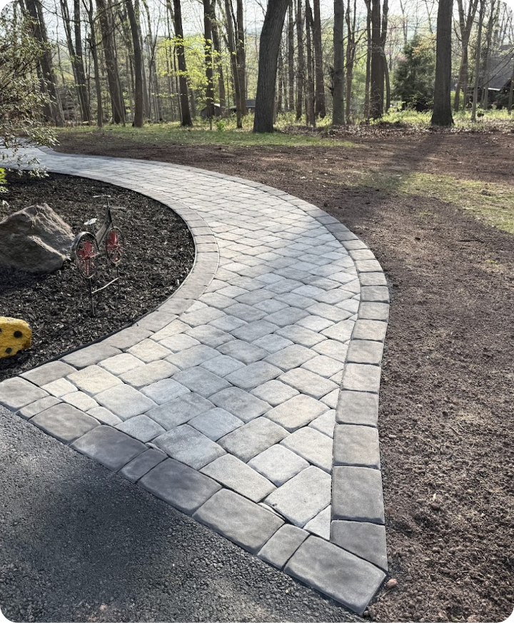 Curved paved pathway in a wooded backyard with trees and some outdoor decorations.