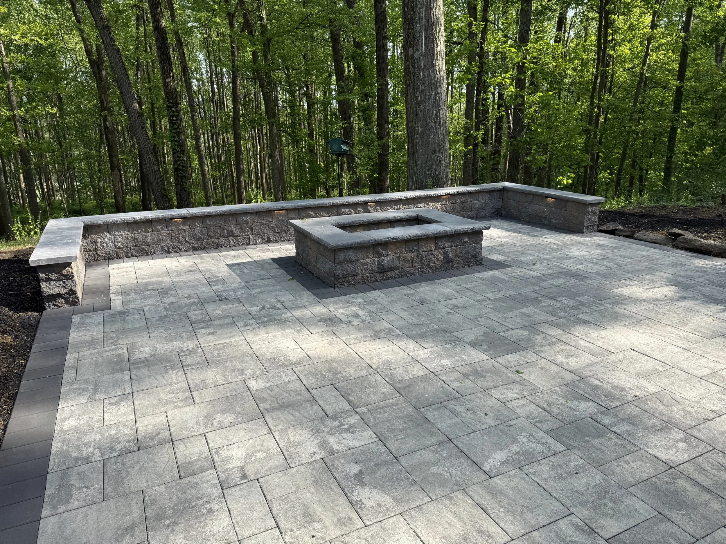 A stone patio with steps in a wooded area with tall trees and green foliage.