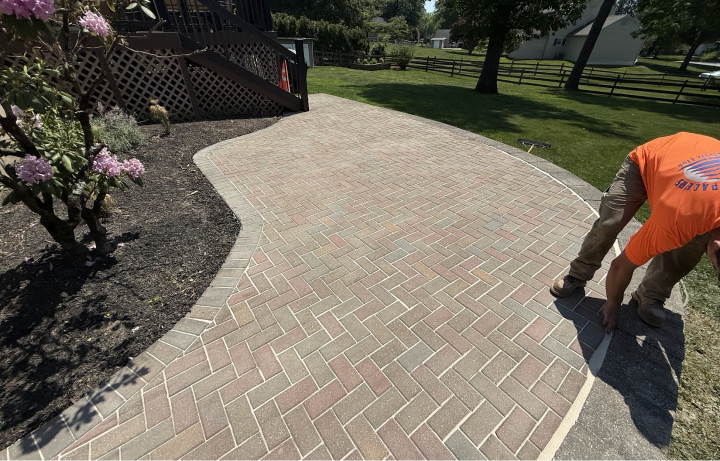 A person installing a border around a brick-paved walkway in a backyard, with a flower bed on the left and a grassy area with trees and a fence in the background.