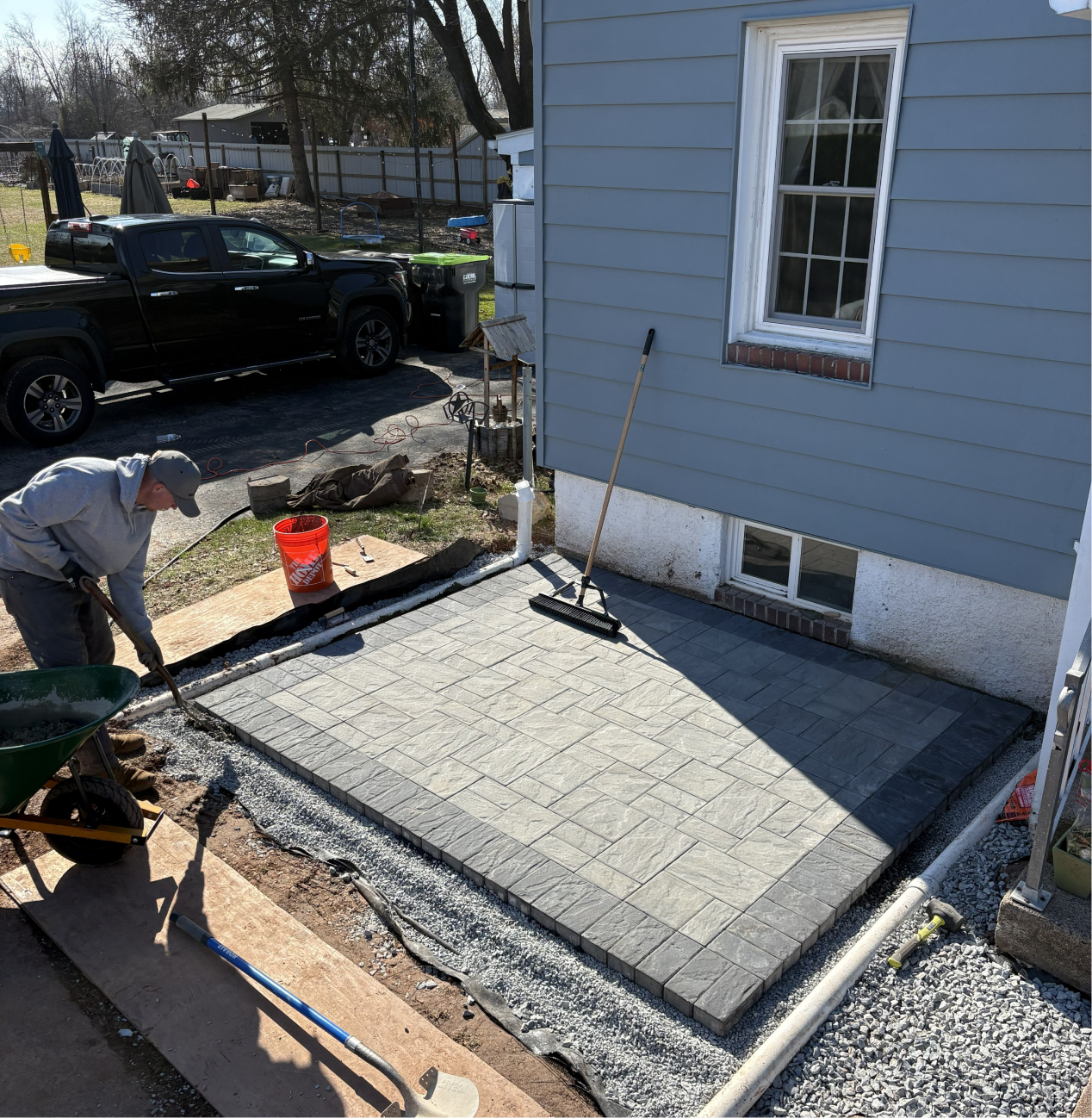 A person working on installing a new concrete patio in a backyard, with paving stones laid out, construction tools, and a wheelbarrow nearby. A black truck is parked in the driveway, and a blue house with white trim is in view.