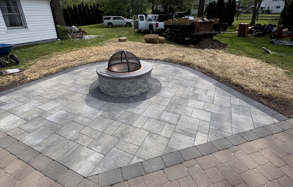 A newly constructed outdoor fire pit area with paving stones, surrounded by a border of brick pavers, and a circular fire pit in the center with a mesh cover. Landscaping in progress with straw laid around the paved area, and a backyard with vehicles and tools in the background.