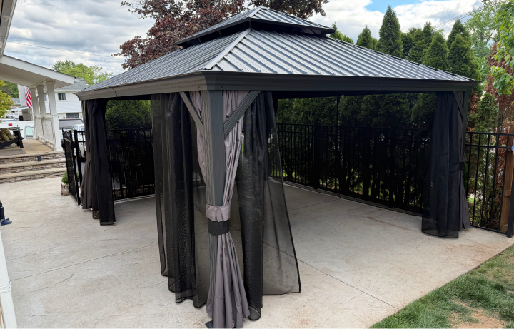 A black metal gazebo with curtains on a concrete patio, surrounded by a black metal fence, with trees and a cloudy sky in the background.