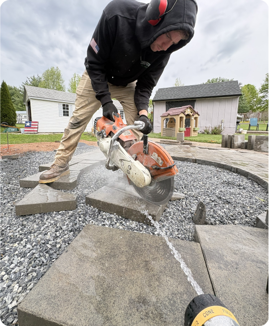A person using a concrete saw to cut a stone paver in an outdoor yard, with houses and trees in the background.