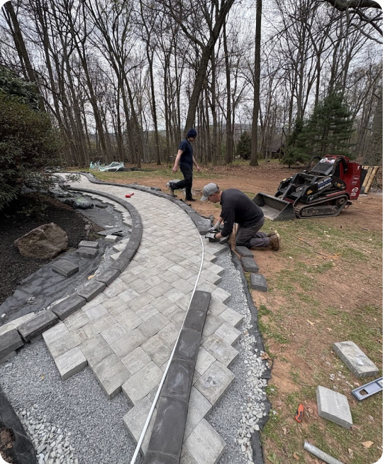 Two workers laying pavers to build a curved walkway in a backyard, with a small skid-steer loader nearby and a wooded area in the background.