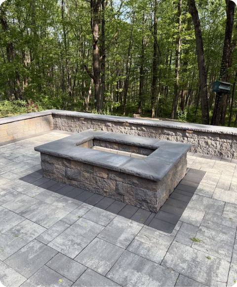 Stone seating area with a fire pit on a paved patio surrounded by trees.