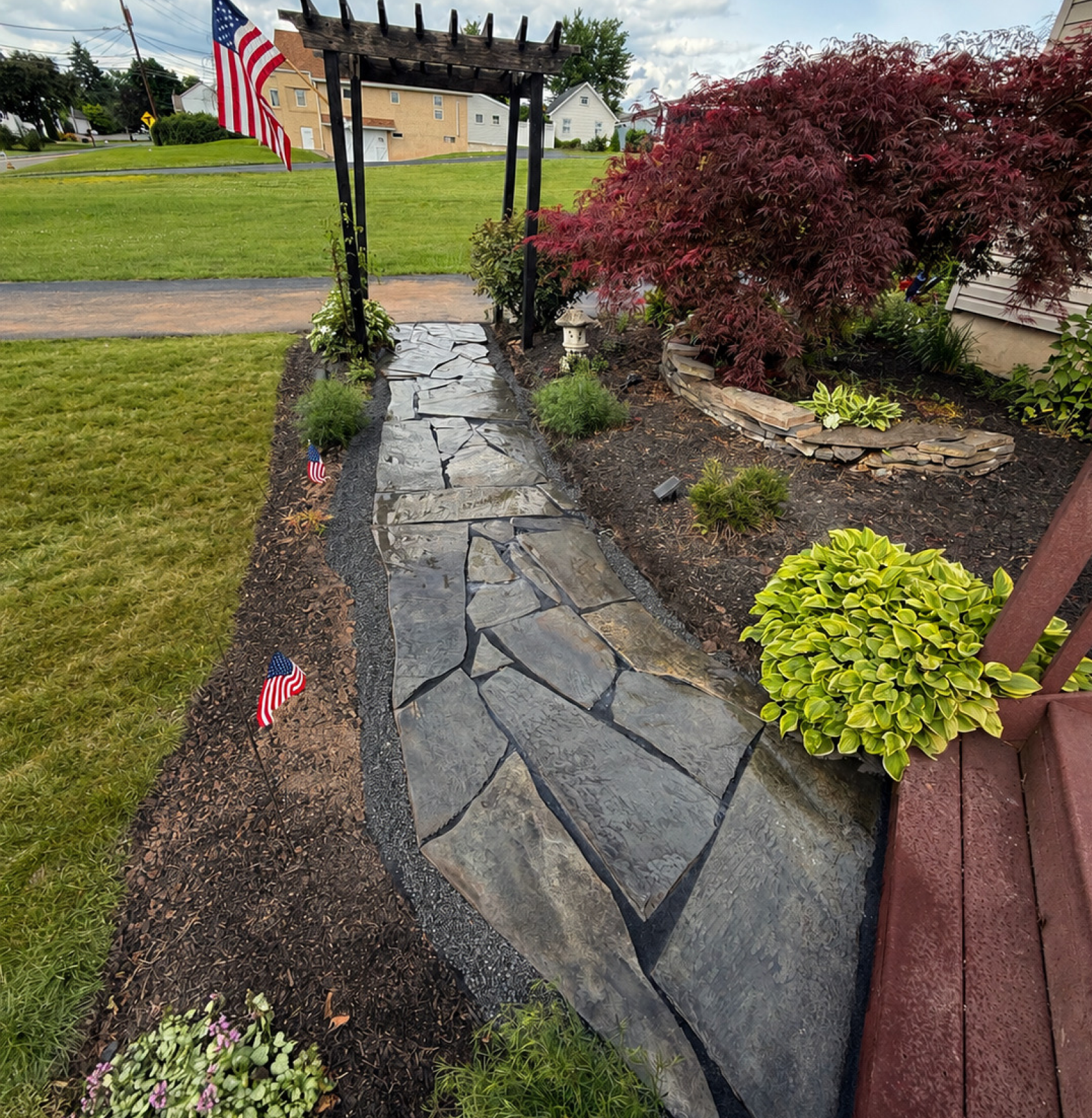 A garden pathway made of large flat stones bordered by dark gravel, leading through a flower bed with a small white decorative lantern, small American flags, green plants, and a large red-leafed shrub, with a house and parked cars in the background.