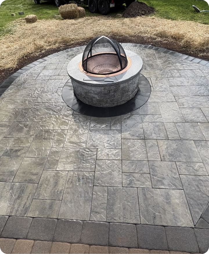 A circular fire pit with a glass screen sits on a newly paved patio with gray textured tiles. There are hay bales and construction materials in the background, indicating ongoing outdoor work.