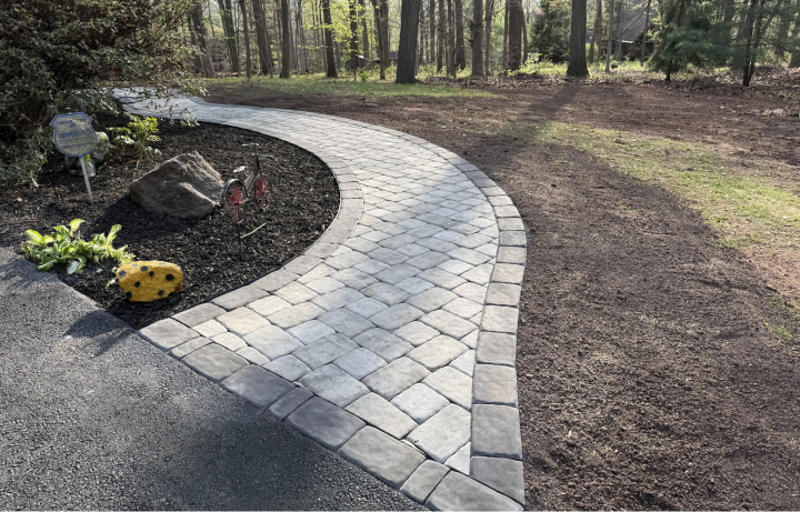 Curved stone pathway in a backyard with a flower bed, decorative rocks, and a wooded area in the background.