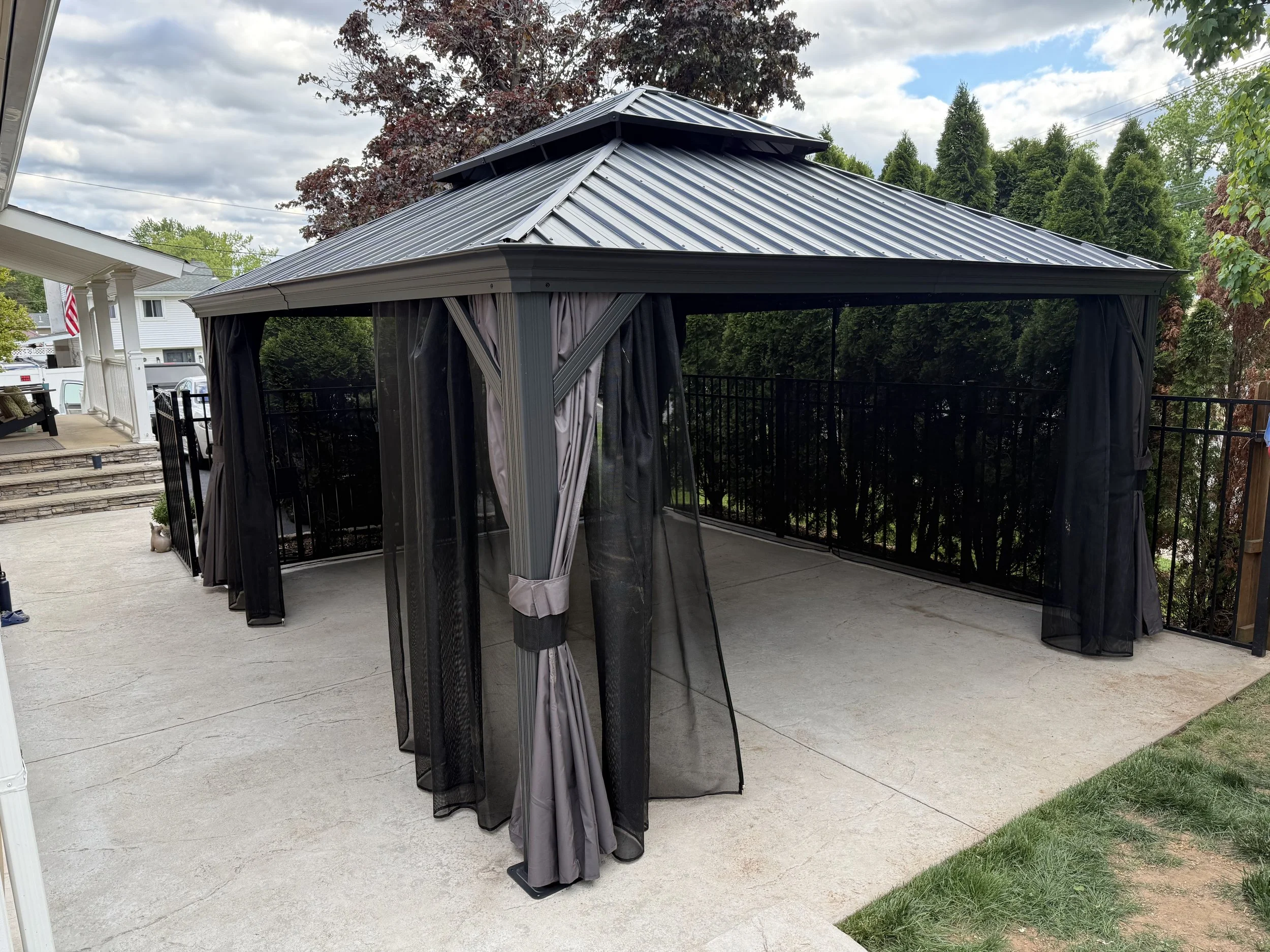 A gray outdoor gazebo with curtains on a concrete patio, surrounded by trees and a black fence, with houses and an American flag visible in the background.