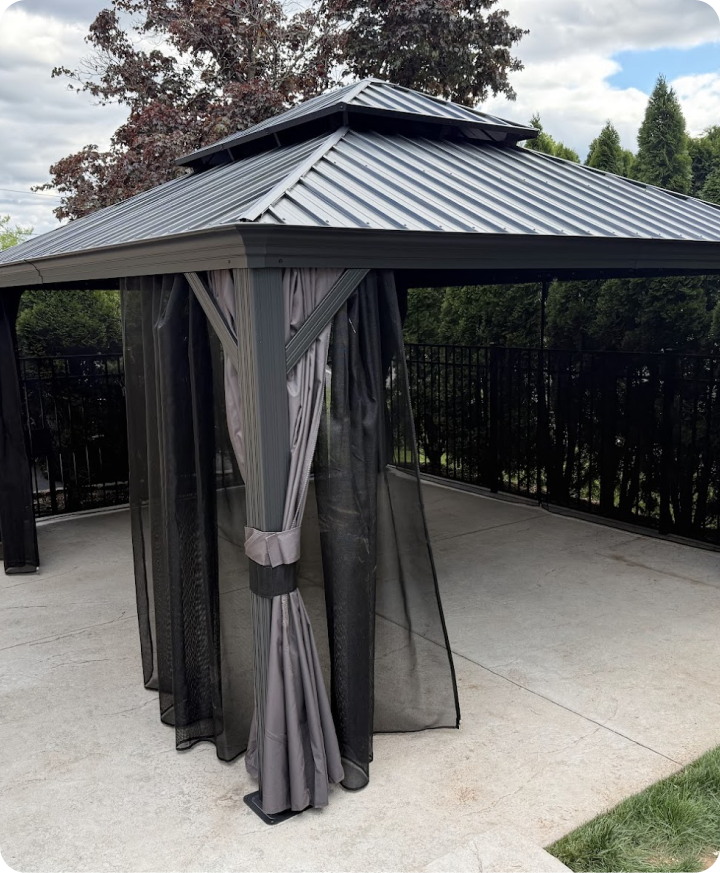 A metal gazebo with a metal roof, black mesh curtains tied back on a concrete patio, surrounded by a black metal fence and green trees in the background.