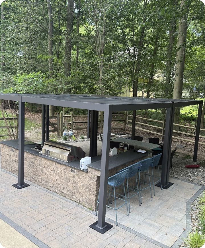 Outdoor kitchen with a stone counter, built-in grill, under a metal pergola, surrounded by trees and a fenced yard.