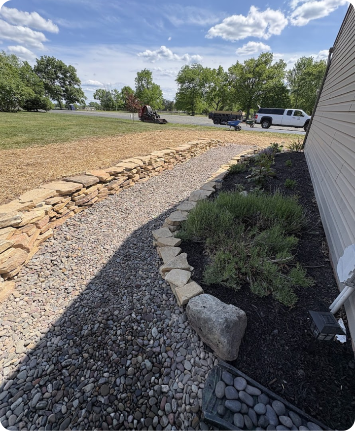 View of a landscaped yard with a gravel pathway bordered by large rocks, small plants, and a white house siding on the right. In the background, a lawn, trees, a road, and parked trucks and equipment are visible under a partly cloudy sky.