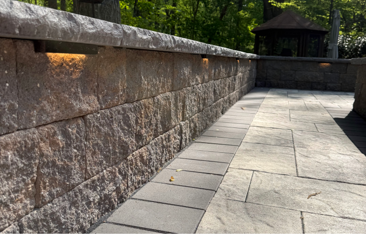 Close-up of a stone retaining wall and paved walkway outdoors, with trees and a small pavilion in the background.