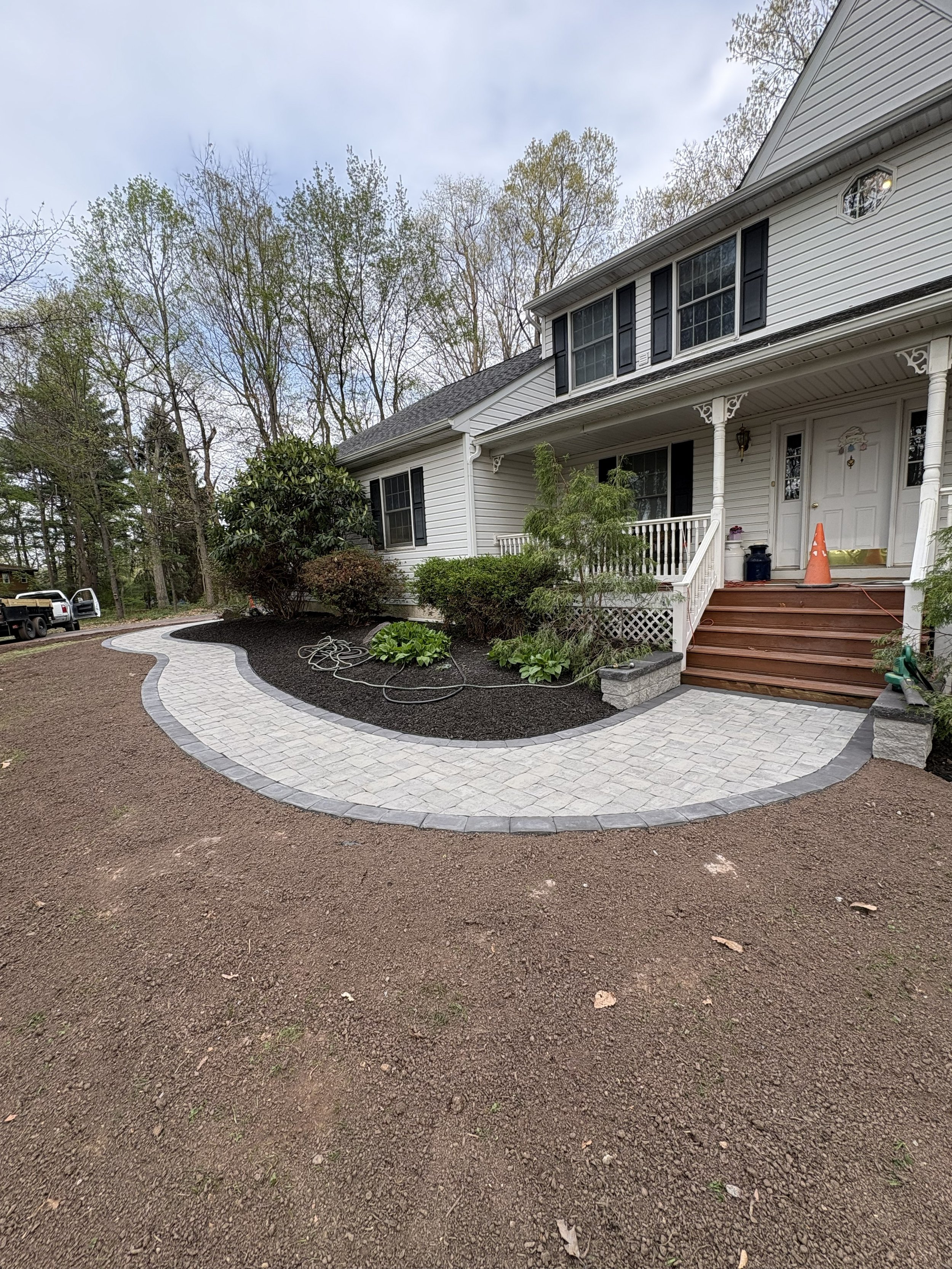 Newly paved circular driveway with a brick border in front of a white house with black shutters and a porch, surrounded by trees and garden beds.