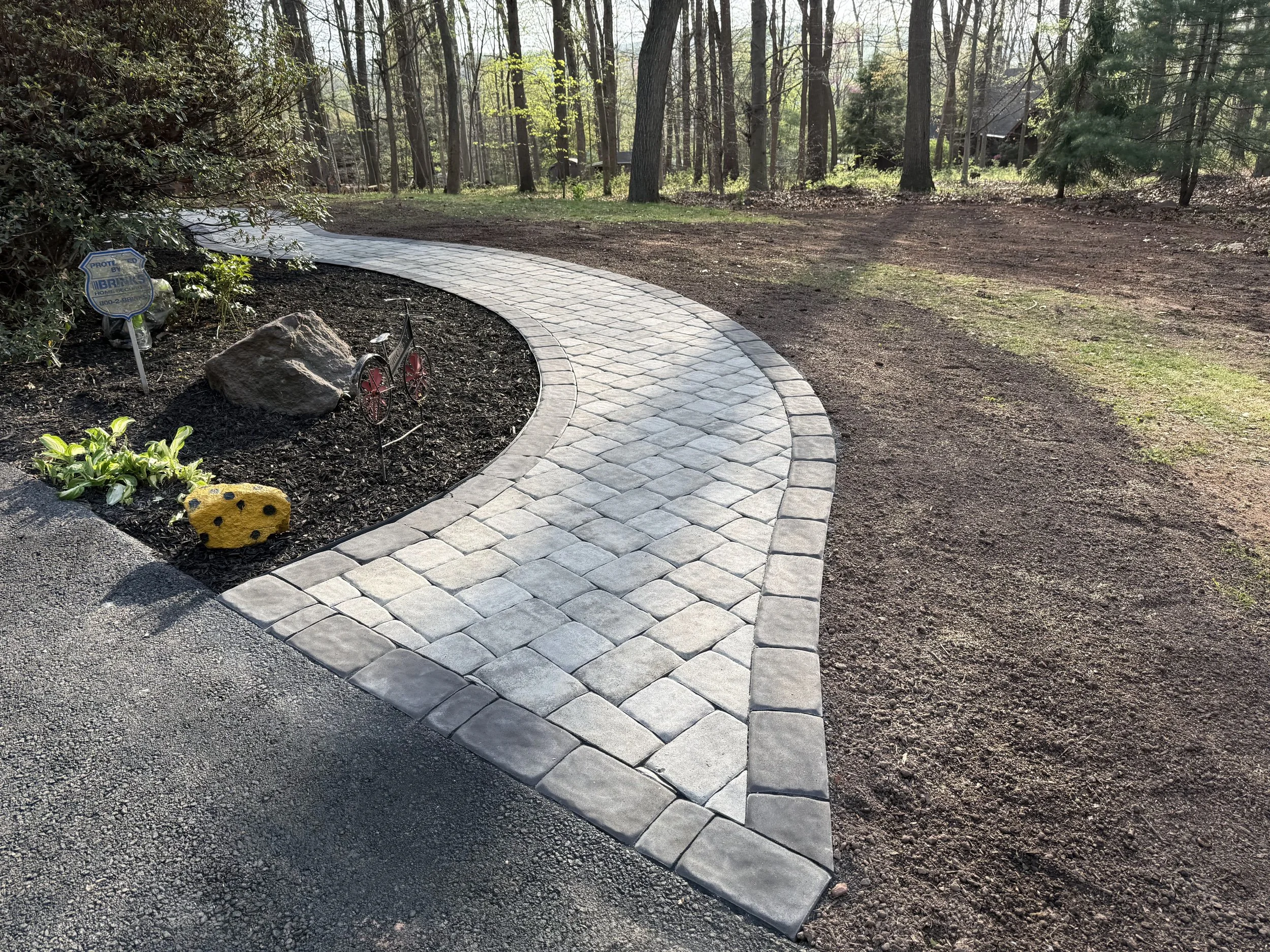 A curved paved walkway in a wooded area with trees, bushes, small plants, and a small yellow rock in the garden bed on the left side.