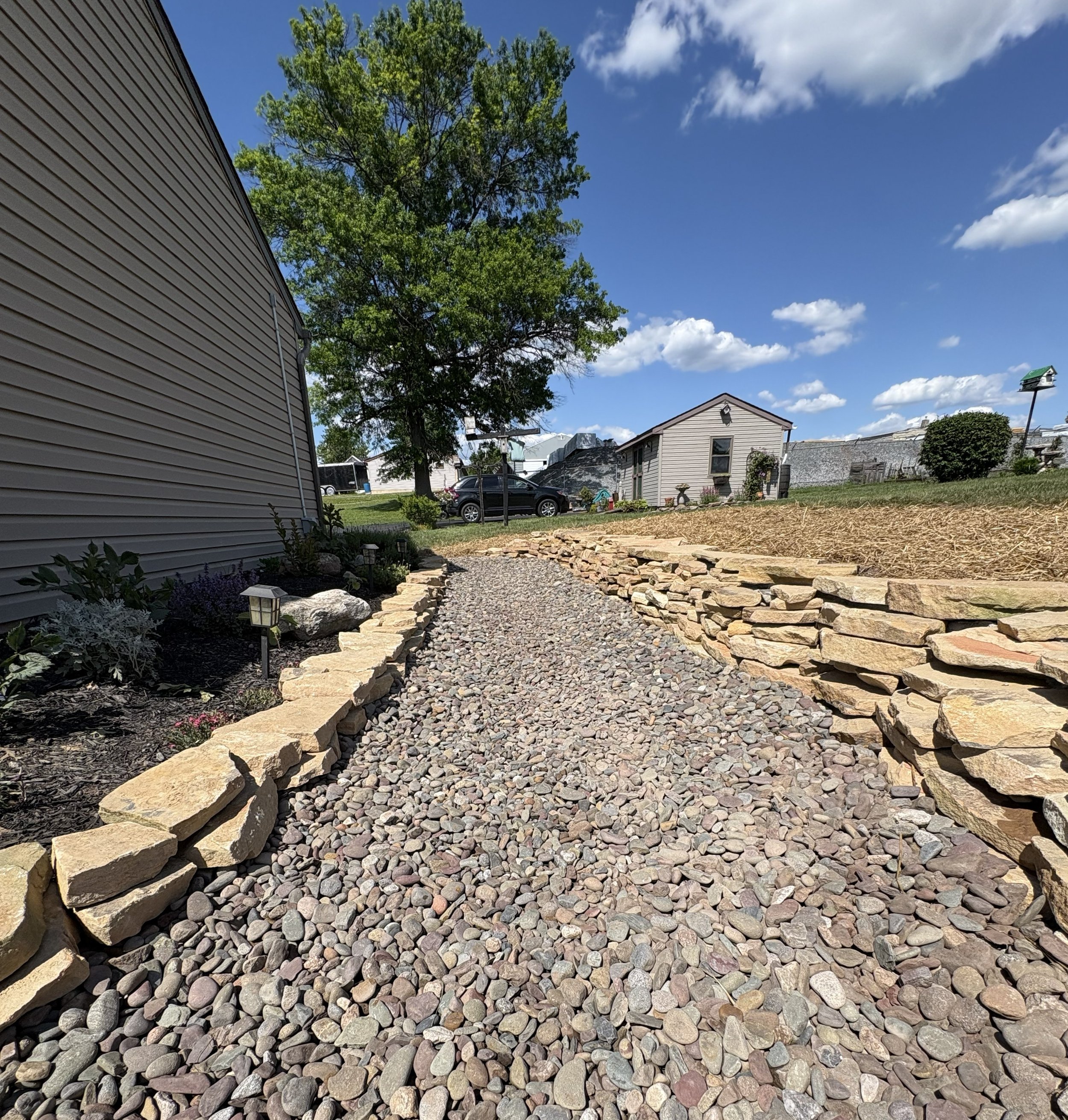 Backyard with a gravel pathway bordered by large flat stones, green bushes, a tall tree with green leaves, a house, and a blue sky with scattered white clouds.