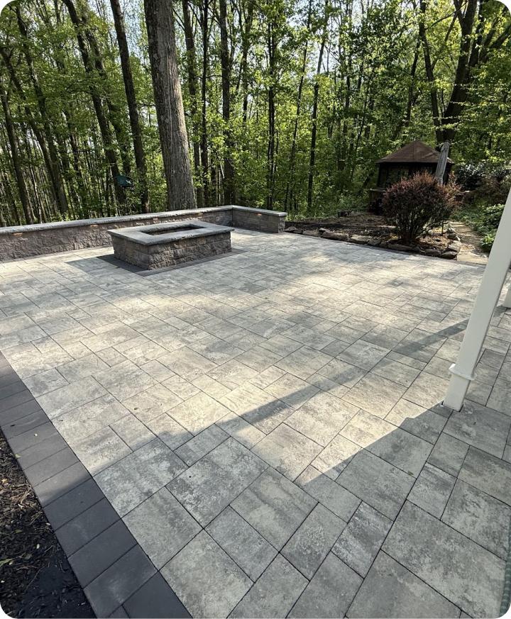 Patio with gray stone pavers, a built-in fire pit, and a wooded backyard with trees and a small tower structure.