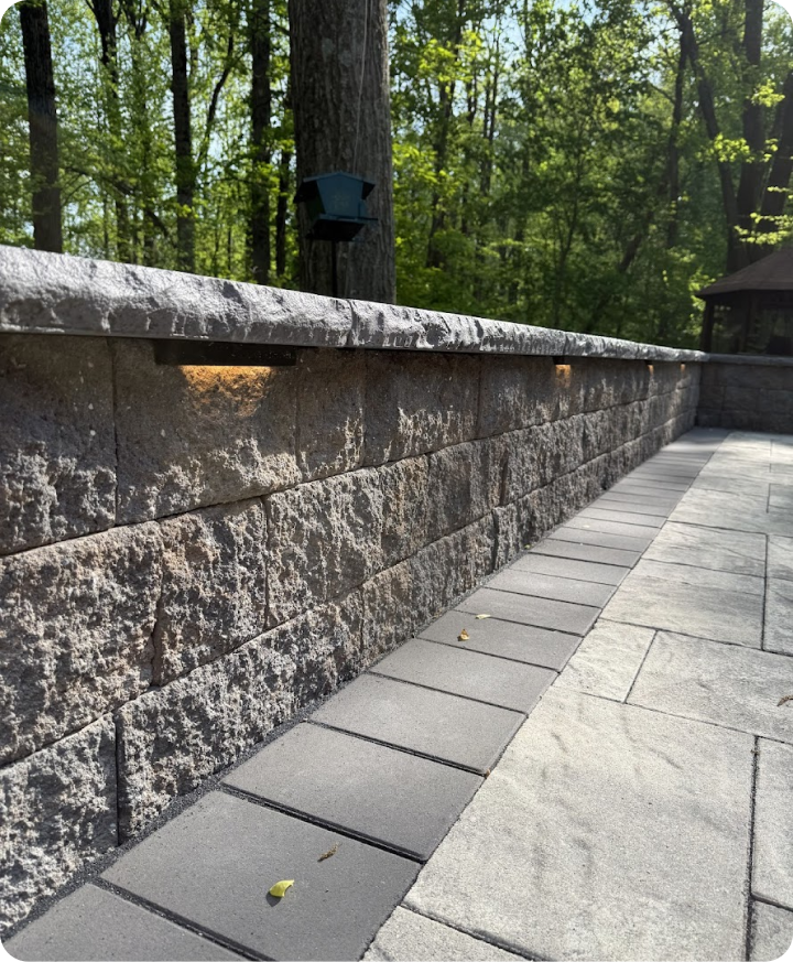 Close-up view of a stone retaining wall and paved outdoor area with trees in the background.