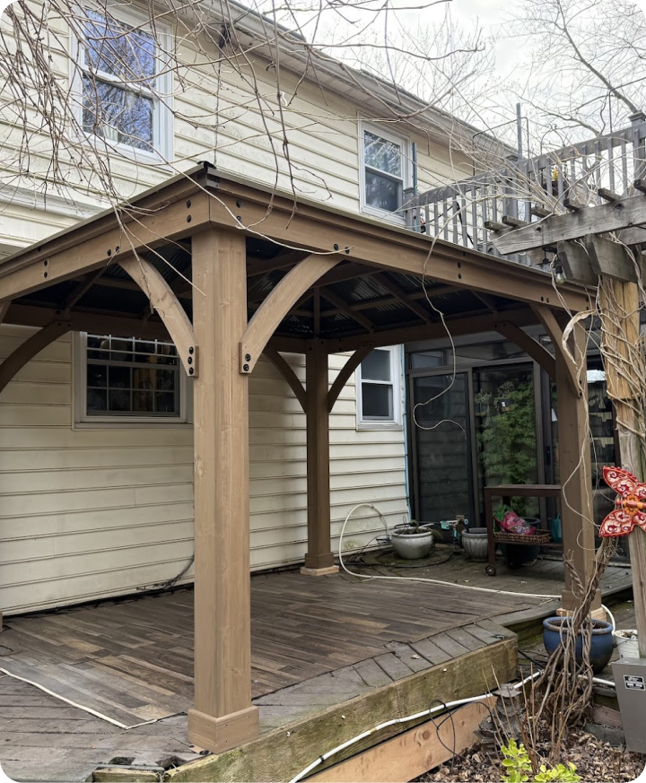 Wooden patio cover with supporting beams and curved braces attached to the back of a beige house with window, over a wooden deck. Tree branches are partly visible in the foreground.