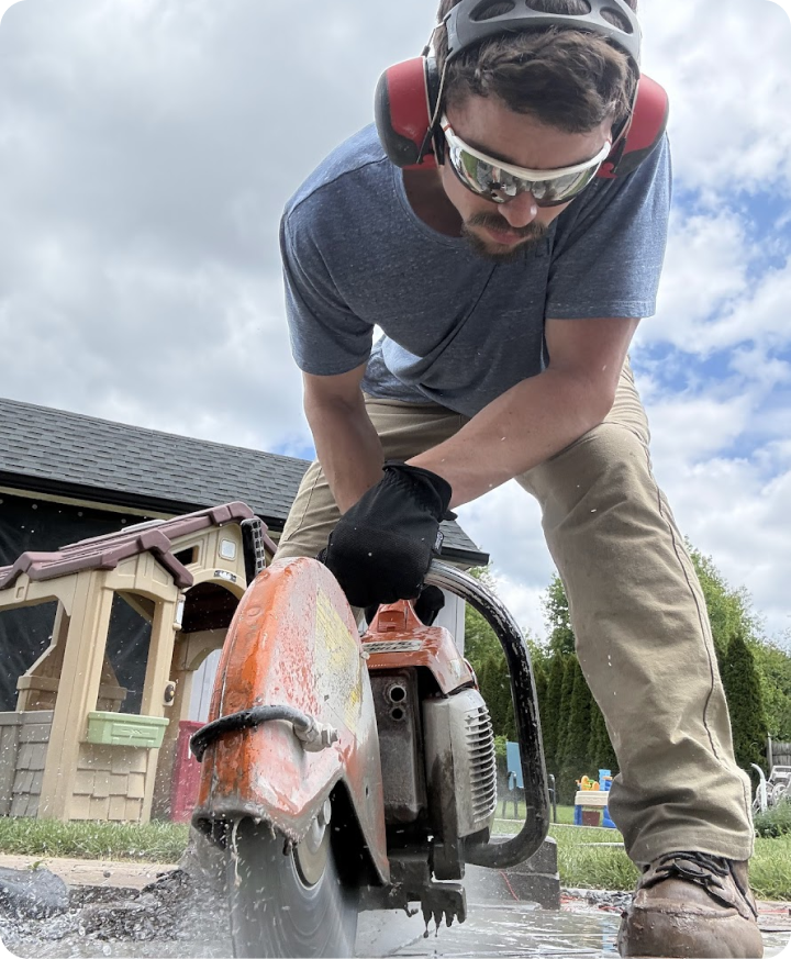A man wearing safety goggles, ear protection, a gray t-shirt, and beige pants operates a cutting saw outdoors on a rainy day, with a playhouse and trees in the background.