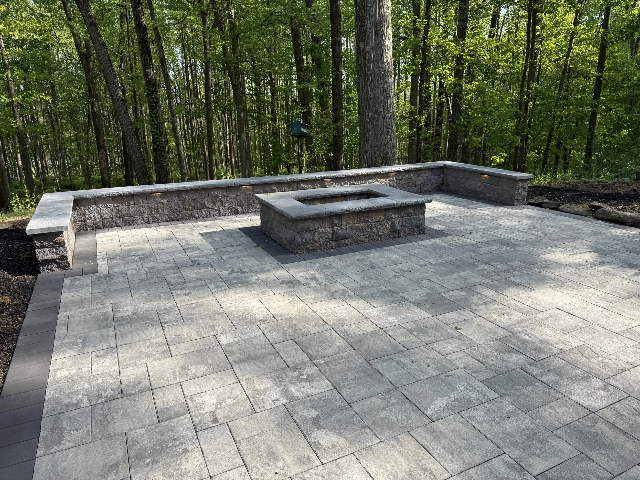 A stone patio with a low stone wall in a wooded area with tall trees and dense green foliage.