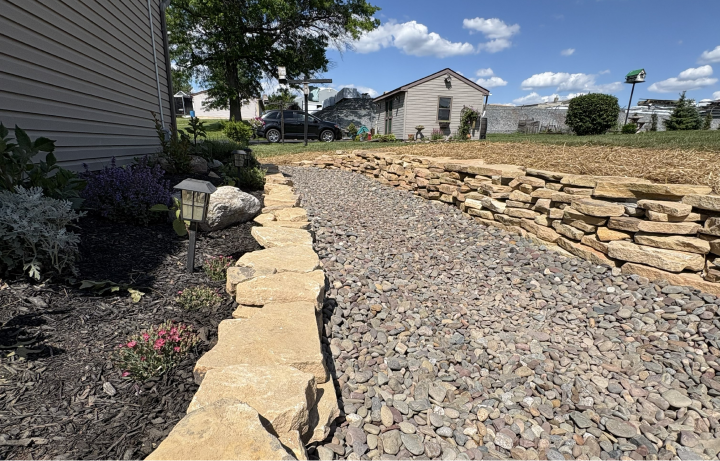 A landscaped backyard with a gravel pathway bordered by stone walls on both sides, a small garden bed with plants and flowers on the left, a lamppost along the path, and a house, trees, and a sunny sky with clouds in the background.