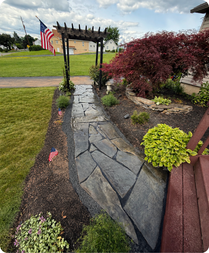 A garden pathway made of large, flat stones bordered by dark gravel, leading to a wooden archway with an American flag, surrounded by various plants and small American flags on stakes.