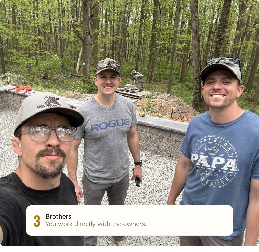 Three men smiling at the camera outdoors in front of a new brick wall, with a forested background and construction tools and materials nearby.
