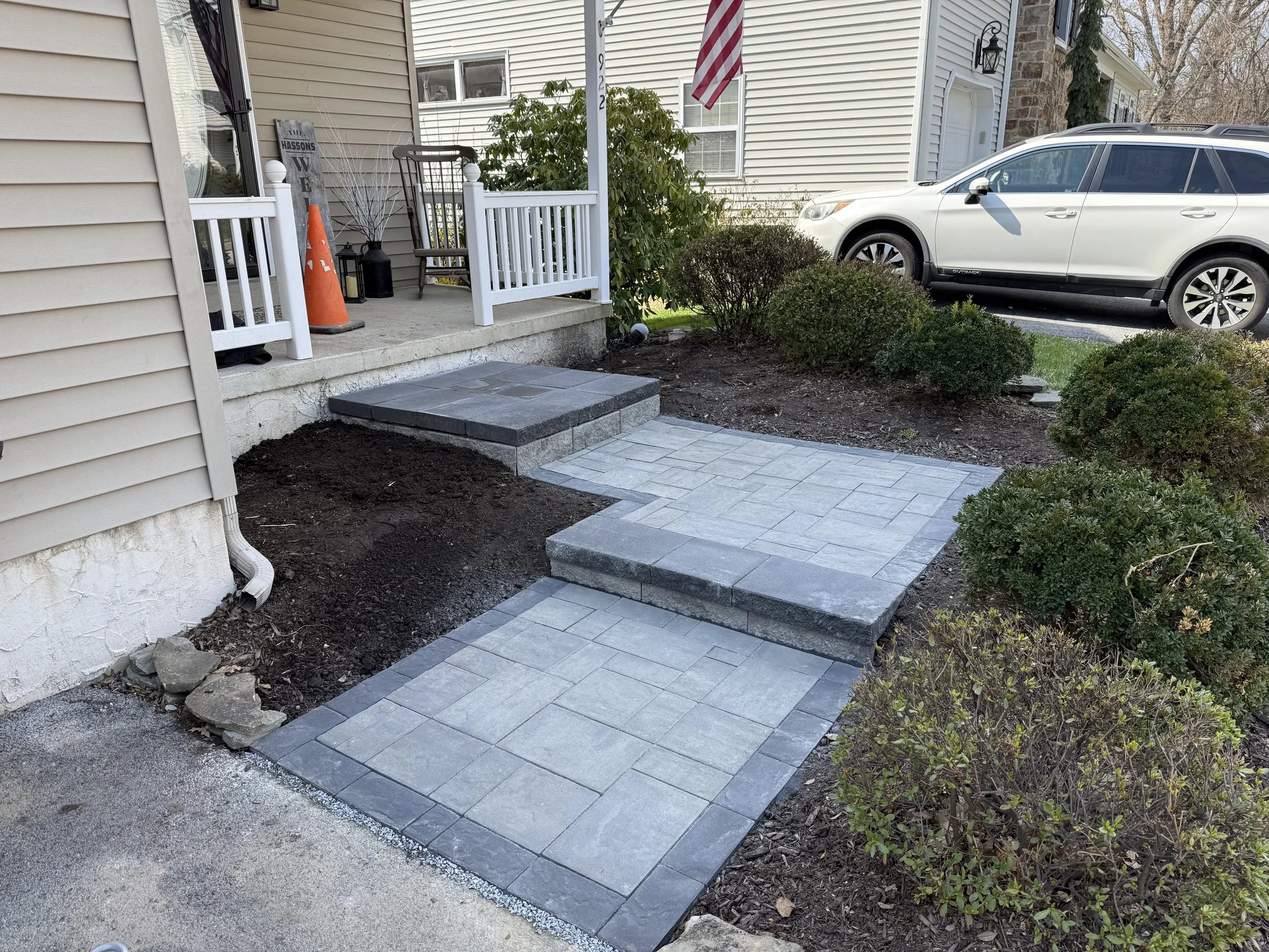 A front porch with a small set of gray steps leading up to a white deck with white railings. There are construction cones, black lanterns, a wooden rocking chair, and a house sign. A small American flag is hanging from a pole. A white SUV is parked i