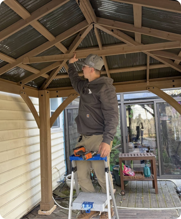 A person installing or inspecting a wooden gazebo structure on a porch, standing on a step ladder with tools attached.