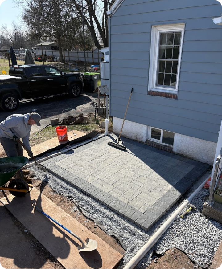 A person working in a yard, laying new paving stones for a walkway beside a blue house with a window. There is a black truck parked in the driveway and gardening tools nearby.