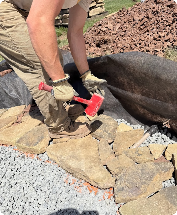 Person placing large rocks to create a border or pathway, using a red hammer, wearing gloves and work boots on a gravel and stone ground, with a pile of rocks and a black fabric liner nearby.