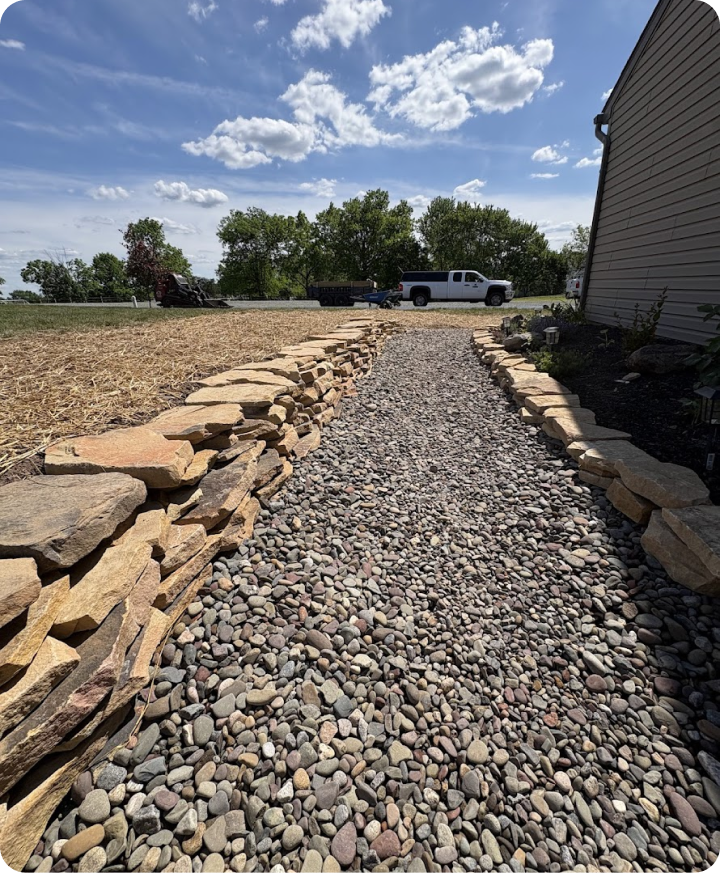 A gravel and stone pathway next to a house on a sunny day with a blue sky and clouds, with trees and vehicles in the background.