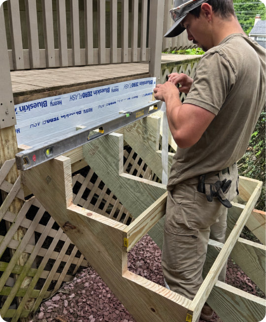 A person working on building a wooden staircase outdoors, using a level and tools, with a deck above and lattice fencing nearby.