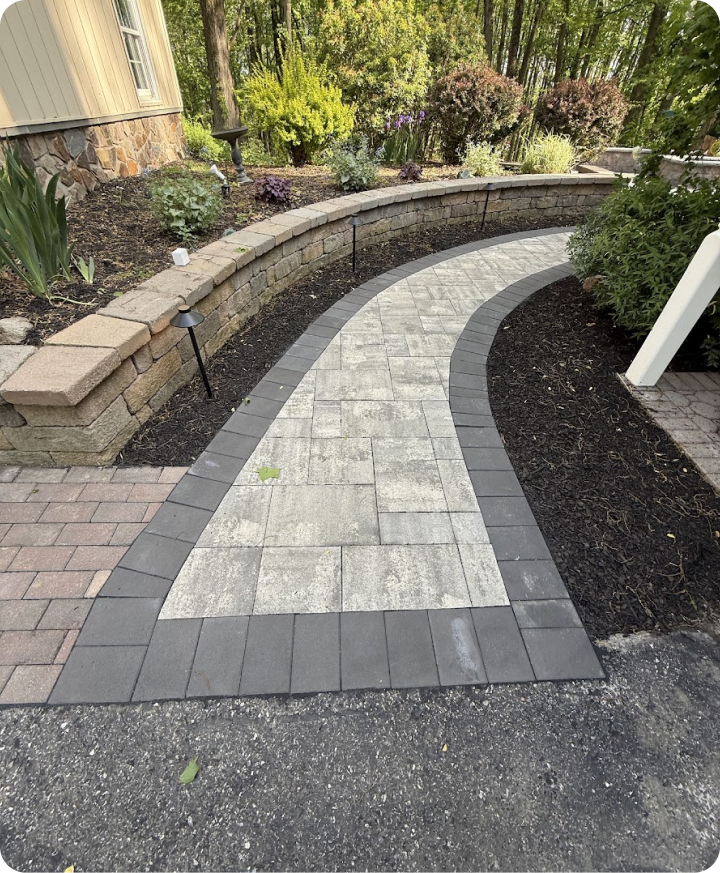 A curved walkway made of gray and beige pavers, bordered by dark gray bricks, with a landscaped garden featuring bushes and trees in the background.