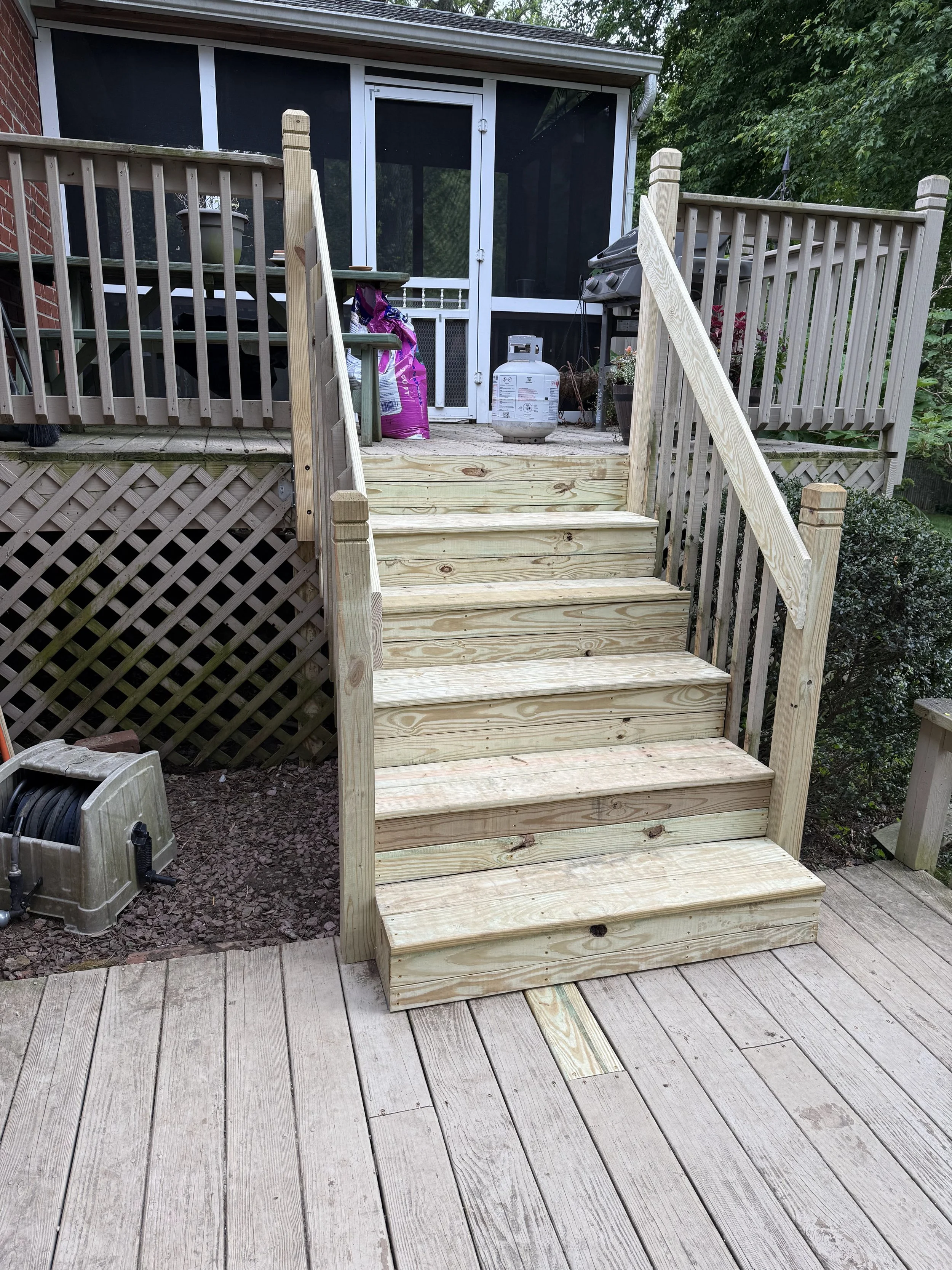 Unfinished wooden deck stairs leading up to a screened porch with black screening and white framing. The deck is attached to a house with brick exterior on the left side. There are plants and trees surrounding the area.