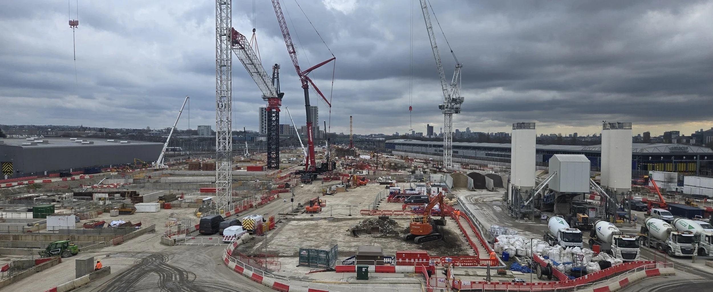 Photo of a large construction site with multiple cranes, construction vehicles, and building materials under a cloudy sky.