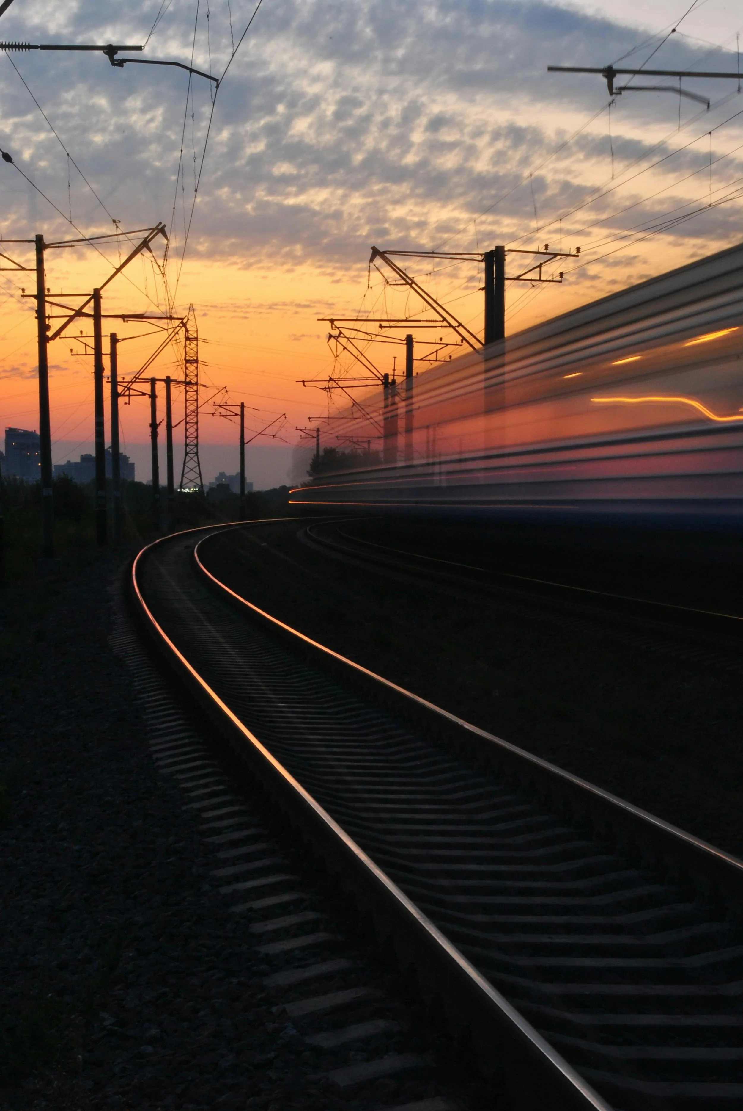 A train moving fast on curved railway tracks during sunset with overhead electric wires and a partly cloudy sky.