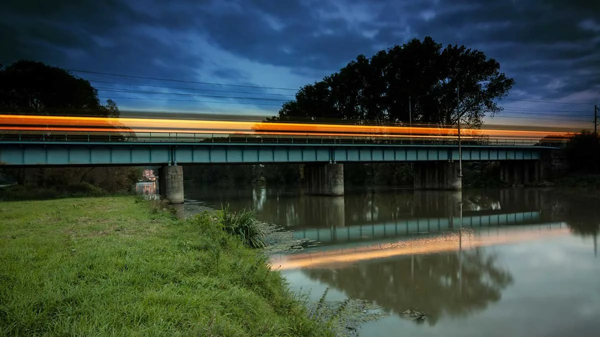 A bridge over a river at dusk with light trails from passing vehicles and their reflections in the water.