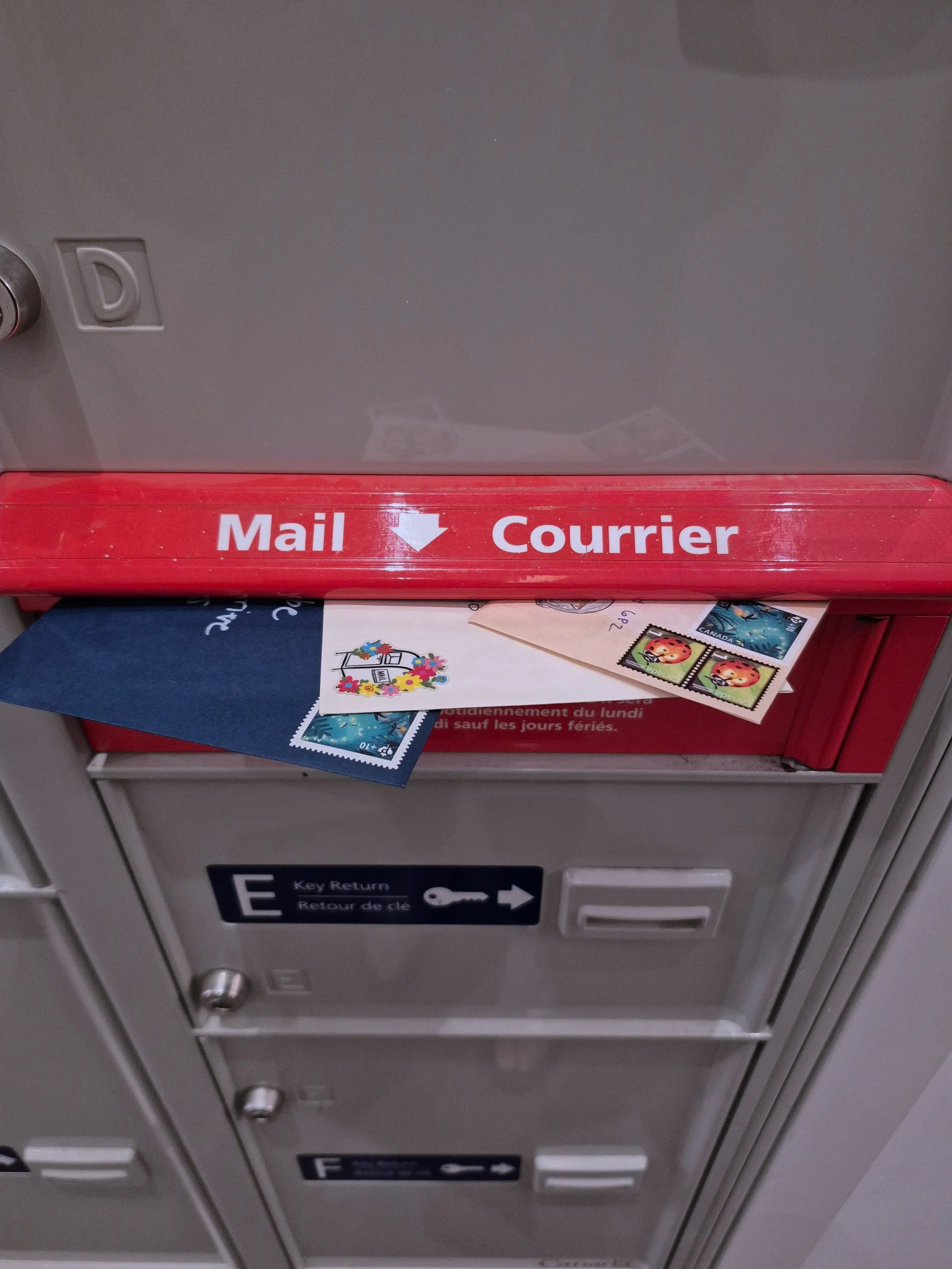 An outgoing mail deposit box with a red sign labeled 'Mail' and 'Courrier' in white letters, with letters and stamps placed inside.